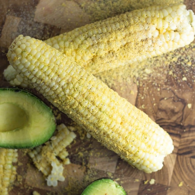 2 ears of corn on the cob covered in nutritional yeast with half an avocado next to them