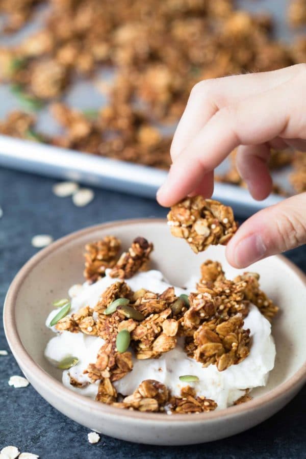 Yogurt in a bowl on a table topped with pumpkin spice granola and a hand holding a granola chunk