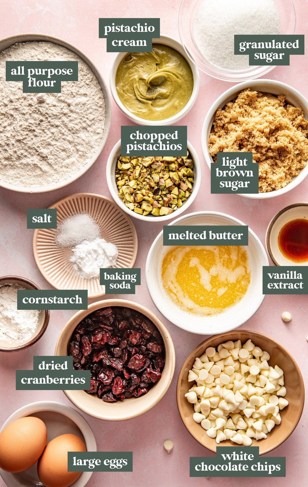 Overhead image showing labeled ingredients for chewy pistachio cranberry cookies, including all-purpose flour, pistachio cream, chopped pistachios, granulated sugar, light brown sugar, melted butter, vanilla extract, white chocolate chips, dried cranberries, large eggs, cornstarch, baking soda, and salt arranged on a pink surface.