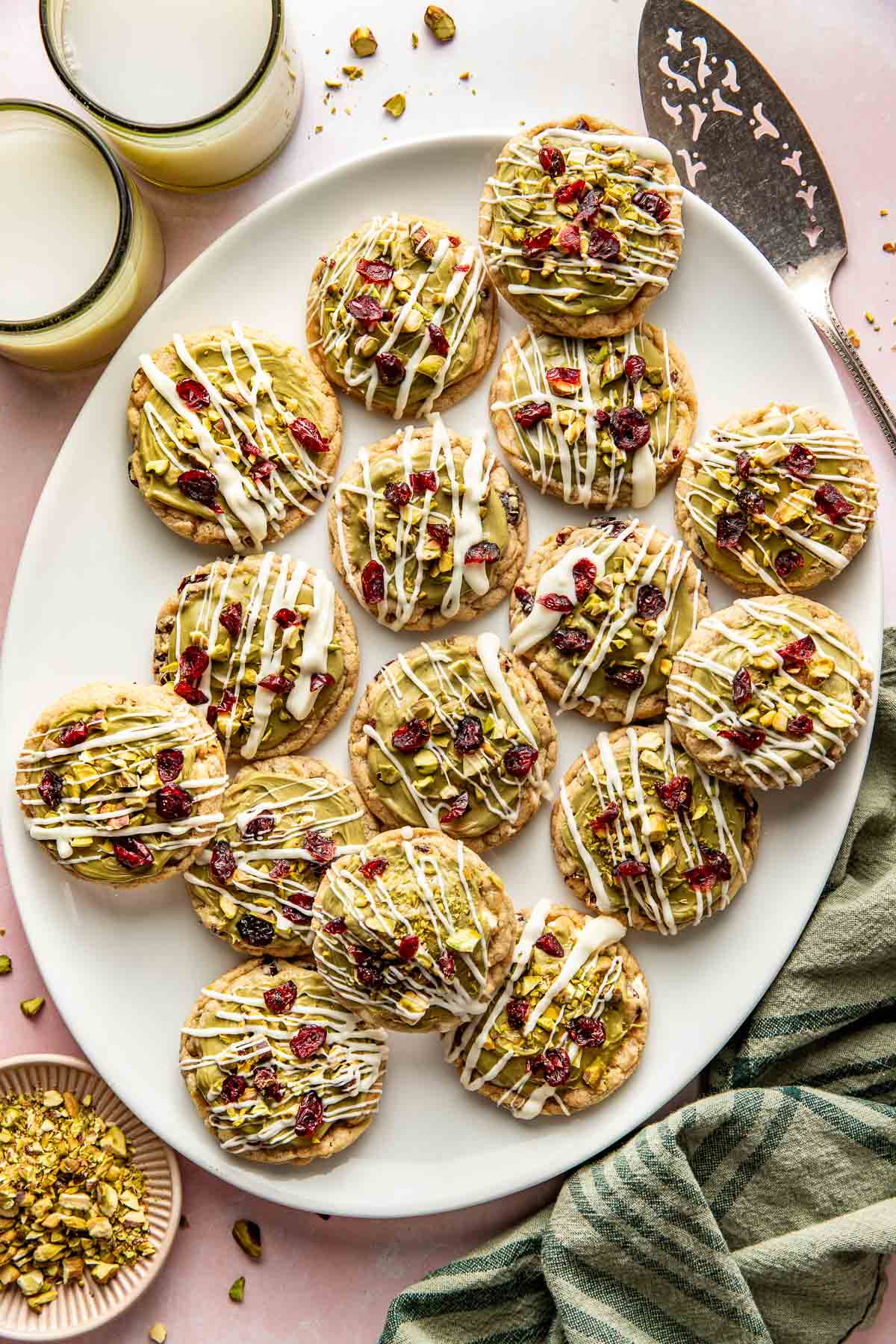 A large white platter filled with pistachio cranberry cookies drizzled with white chocolate and topped with chopped pistachios and cranberries, next to glasses of milk and a green towel.