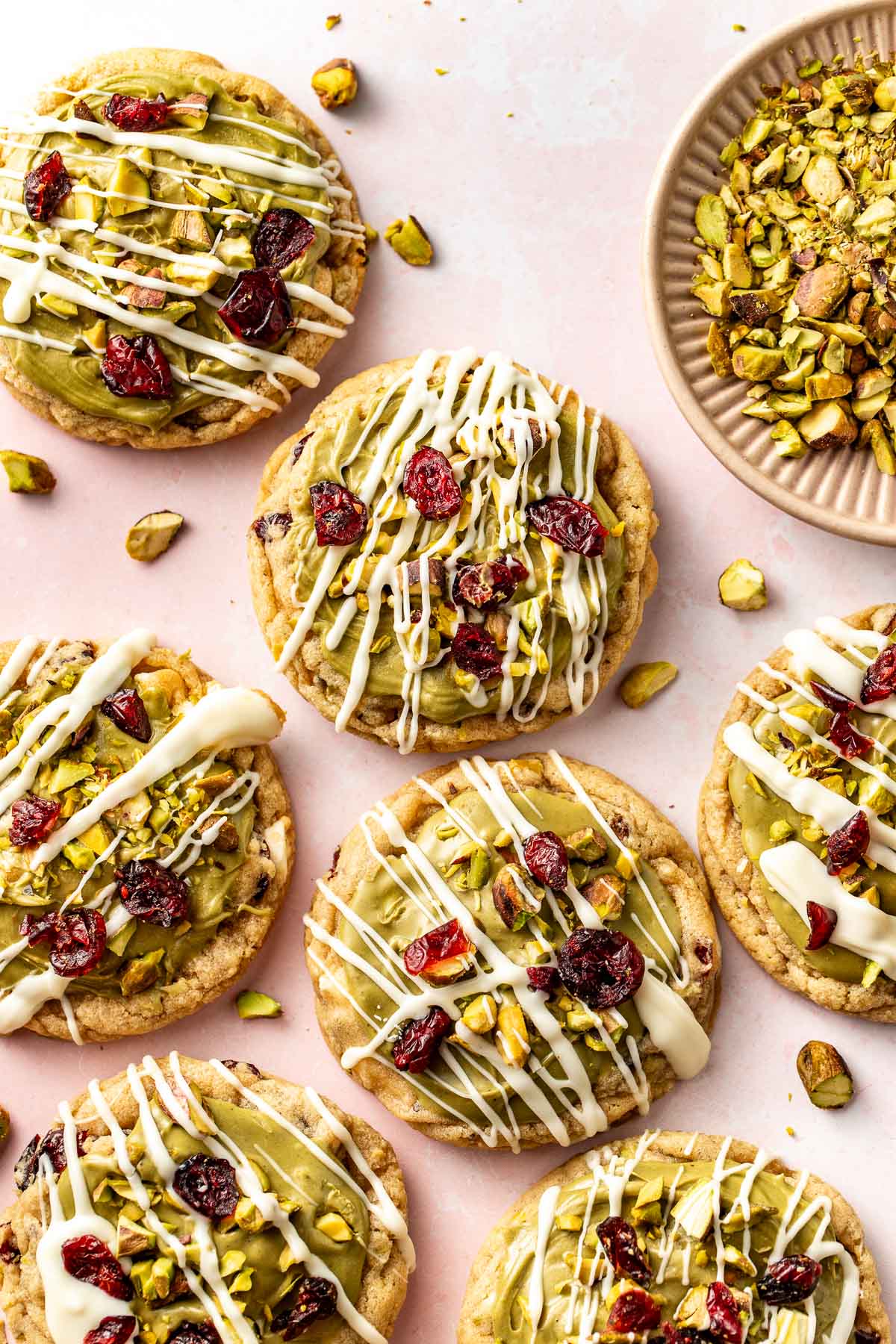 Close-up of pistachio cranberry cookies on a pink surface, decorated with green frosting, white chocolate drizzle, chopped pistachios, and dried cranberries.
