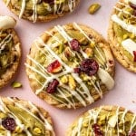 Close-up of pistachio cranberry cookies with white chocolate drizzle and bright red cranberries, surrounded by scattered pistachio pieces.