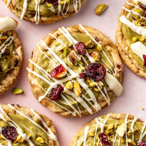 Close-up of pistachio cranberry cookies with white chocolate drizzle and bright red cranberries, surrounded by scattered pistachio pieces.