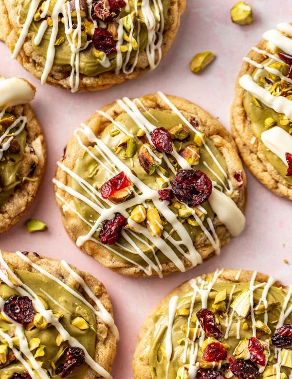 Close-up of pistachio cranberry cookies with white chocolate drizzle and bright red cranberries, surrounded by scattered pistachio pieces.