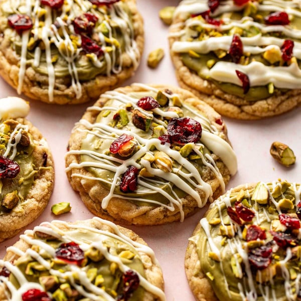Stack of pistachio cranberry cookies showing a soft, chewy interior with white chocolate chips, pistachios, and cranberries, topped with green frosting and drizzled white chocolate.