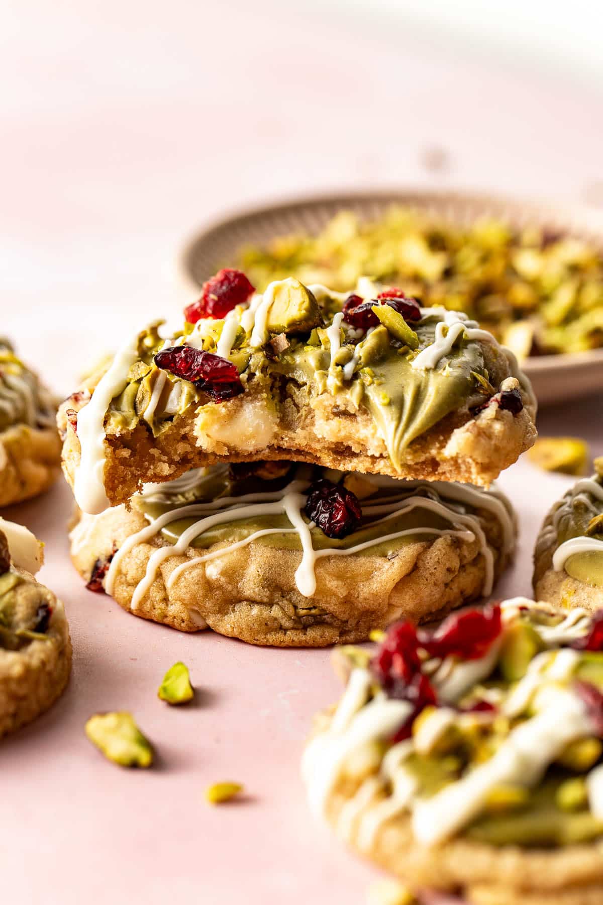 Close-up of a hand holding a pistachio cranberry cookie with a bite taken out, showing its chewy texture and toppings of pistachios, cranberries, and white chocolate drizzle.