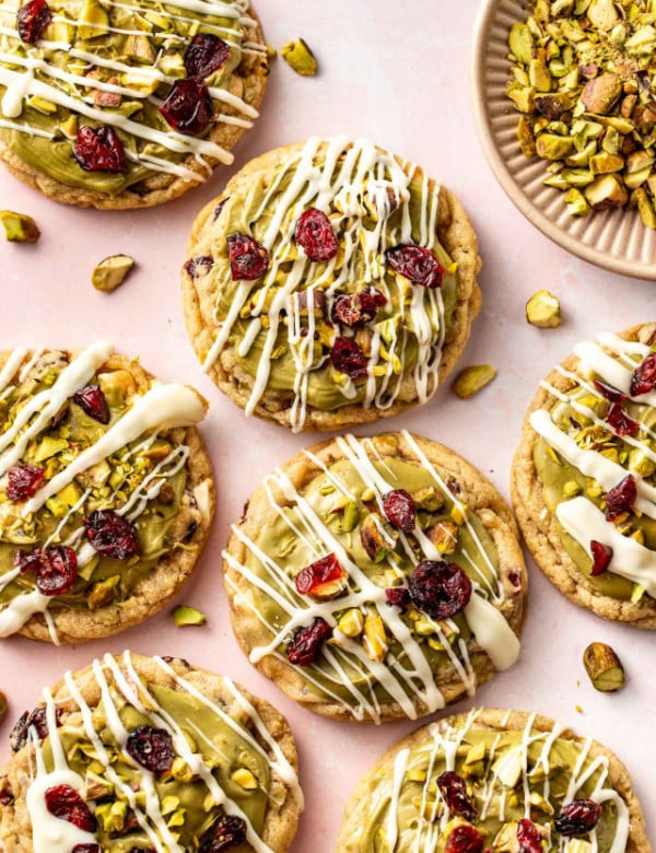Close-up of pistachio cranberry cookies on a pink surface, decorated with green frosting, white chocolate drizzle, chopped pistachios, and dried cranberries.