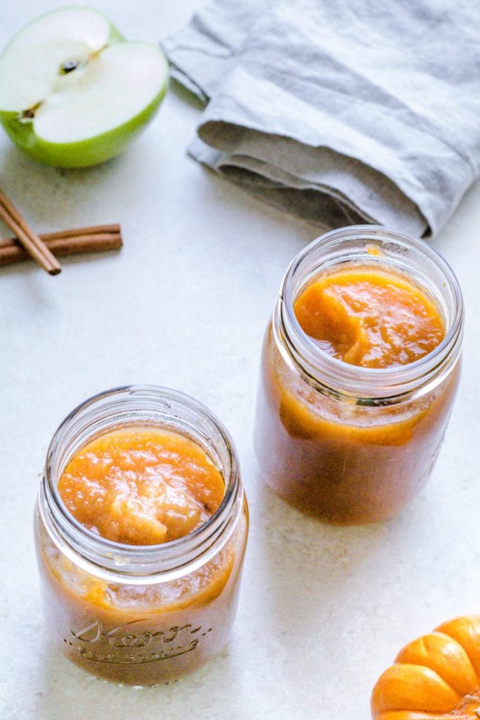 Applesauce made with pumpkin in mason jars on a table. Cinnamon sticks, a green apple and a napkin in the background.