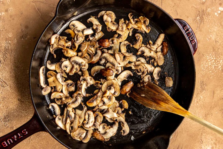 black cast iron skillet with sliced mushrooms being sauteed with a wooden spoon