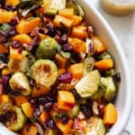 roasted brussels sprouts and butternut squash in a white baking dish garnished with chopped pecans and dried cranberries