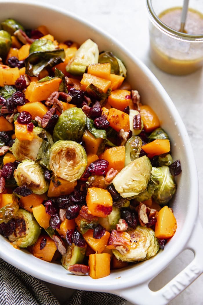 roasted brussels sprouts and butternut squash in a white baking dish garnished with chopped pecans and dried cranberries