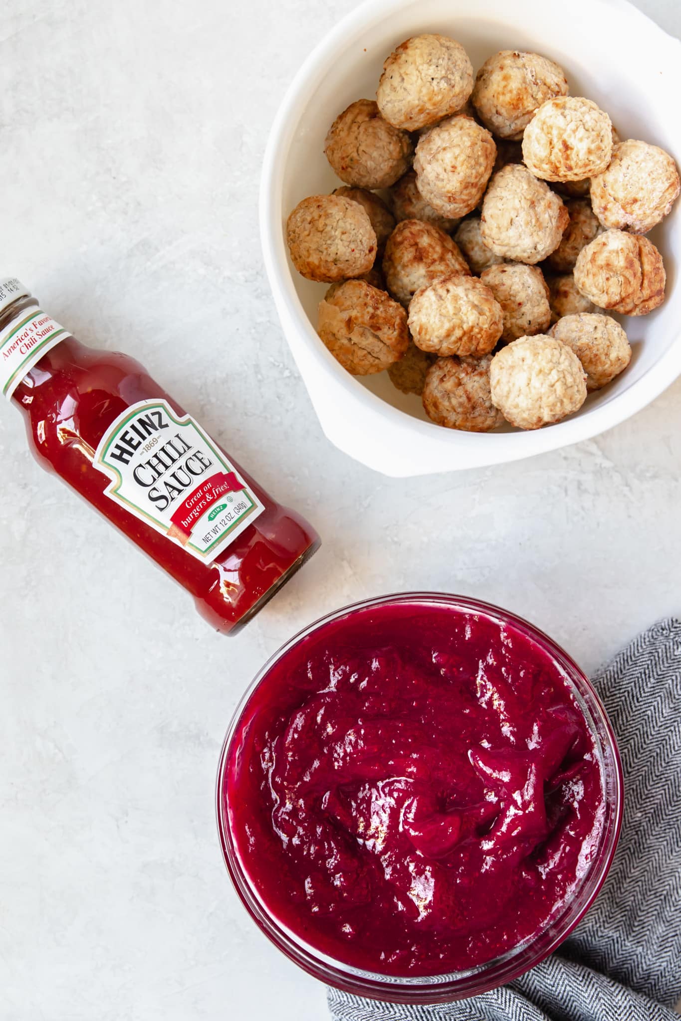 cranberry sauce in a glass bowl, a bottle of chili sauce and frozen meatballs in a white bowl