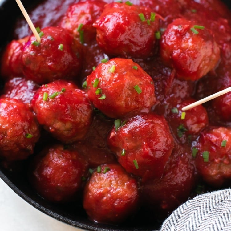 meatballs covered in cranberry sauce and garnished with fresh chives in a cast iron skillet with toothpicks