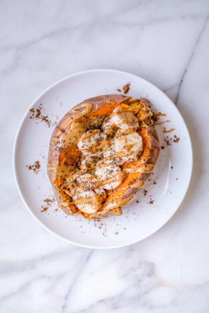 A sweet potato on a white plate filled with banana slices, peanut butter, chia seeds and cinnamon.