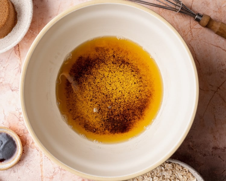 Browned butter in a large mixing bowl with cinnamon and nutmeg sprinkled over the top, shown from an overhead angle on a light countertop with baking tools nearby.