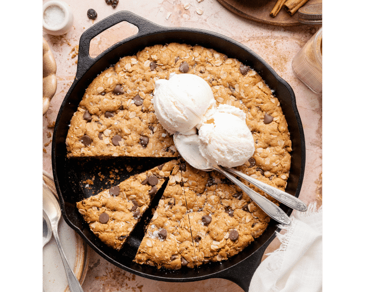An overhead view of an oatmeal raisin chocolate chip cookie skillet baked in a cast iron pan, with a slice removed and two scoops of vanilla ice cream melting on top.