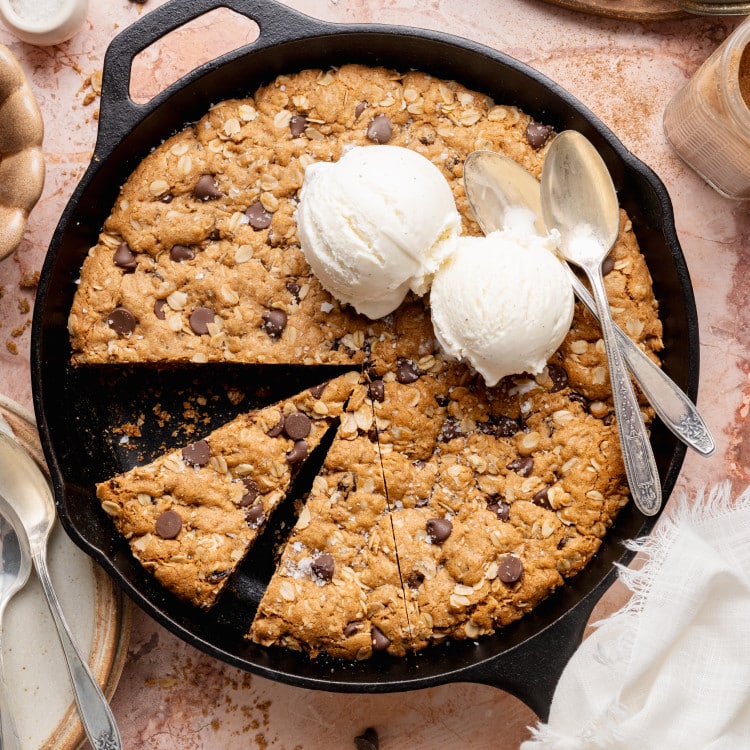 An overhead view of an oatmeal raisin chocolate chip cookie skillet baked in a black cast iron pan, with a slice cut out and two scoops of vanilla ice cream melting on top, surrounded by cinnamon sticks, oats, chocolate chips, and baking props on a neutral surface.
