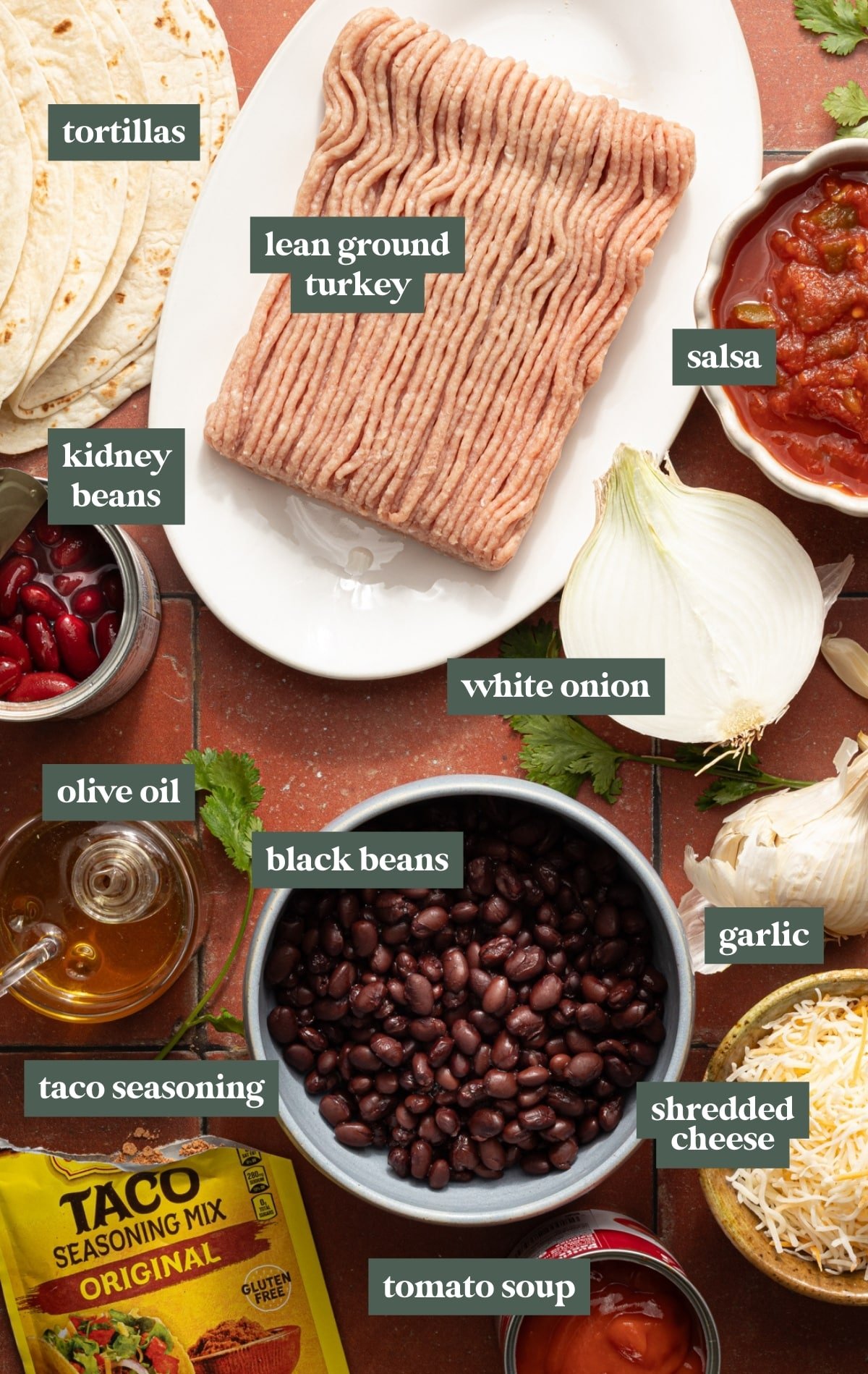 Overhead shot of taco skillet ingredients including tortillas, ground turkey, salsa, beans, onion, garlic, shredded cheese, taco seasoning, tomato soup, and olive oil arranged on a table.