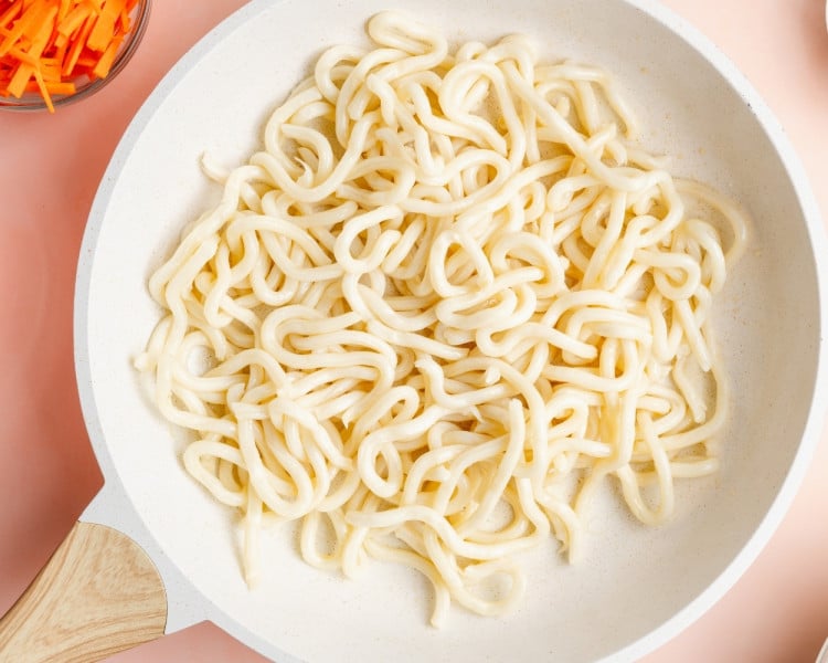 Udon noodles cooking in a skillet with a small amount of water, showing the noodles loosening and becoming pliable.