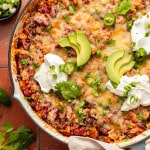 Overhead view of a cheesy taco skillet topped with avocado slices, sour cream, jalapeños, cilantro, and green onions, served with tortillas on the side.