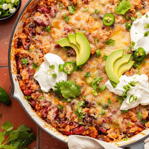 Overhead view of a cheesy taco skillet topped with avocado slices, sour cream, jalapeños, cilantro, and green onions, served with tortillas on the side.