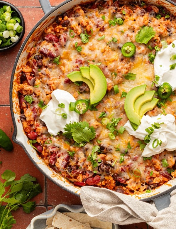 Overhead view of a cheesy taco skillet topped with avocado slices, sour cream, jalapeños, cilantro, and green onions, served with tortillas on the side.