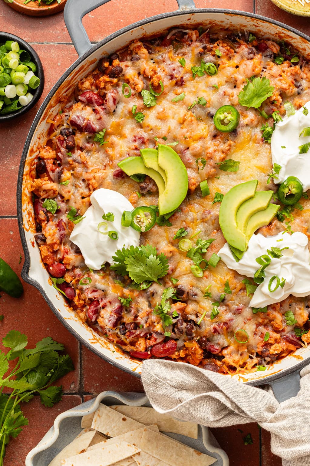 Overhead view of a cheesy taco skillet topped with avocado slices, sour cream, jalapeños, cilantro, and green onions, served with tortillas on the side.