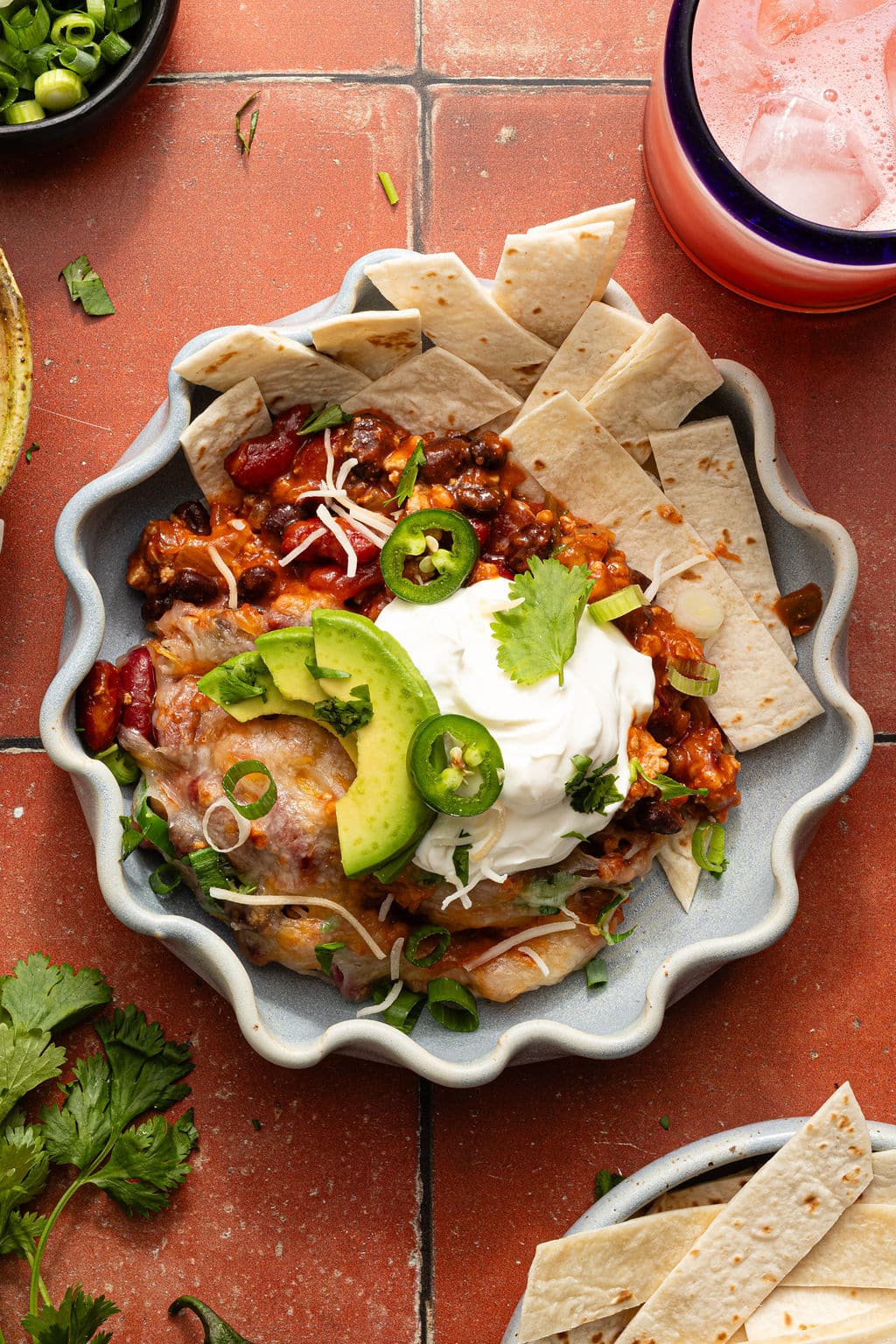 Serving of taco skillet in a scalloped dish topped with avocado, sour cream, jalapeño, and cilantro, with tortilla strips on the side.