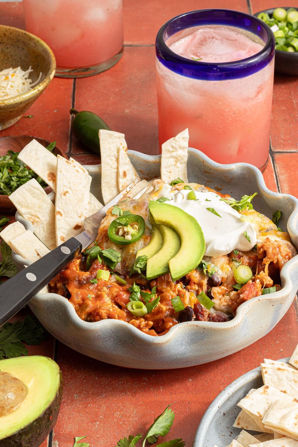 Bowl of taco skillet garnished with avocado, sour cream, and jalapeños, with tortilla strips arranged around the edges and pink drinks in the background.