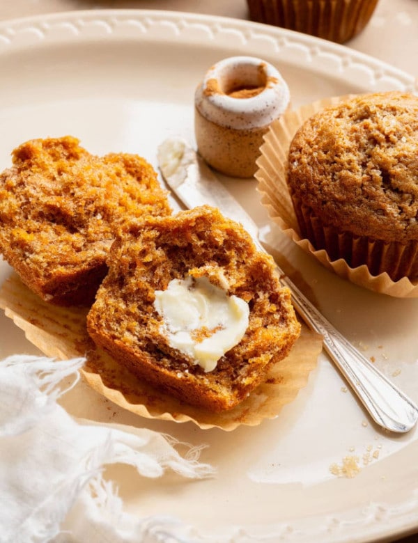 Carrot cake muffin broken in half and topped with softened butter on a parchment liner, with a butter knife and cinnamon jar nearby.