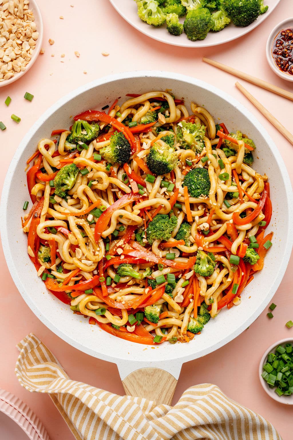 Overhead image of a skillet filled with sesame udon noodles tossed with broccoli, sliced red bell peppers, carrots, and green onions, garnished with chopped peanuts. Small bowls of chili crisp, broccoli, and peanuts surround the pan on a light surface.