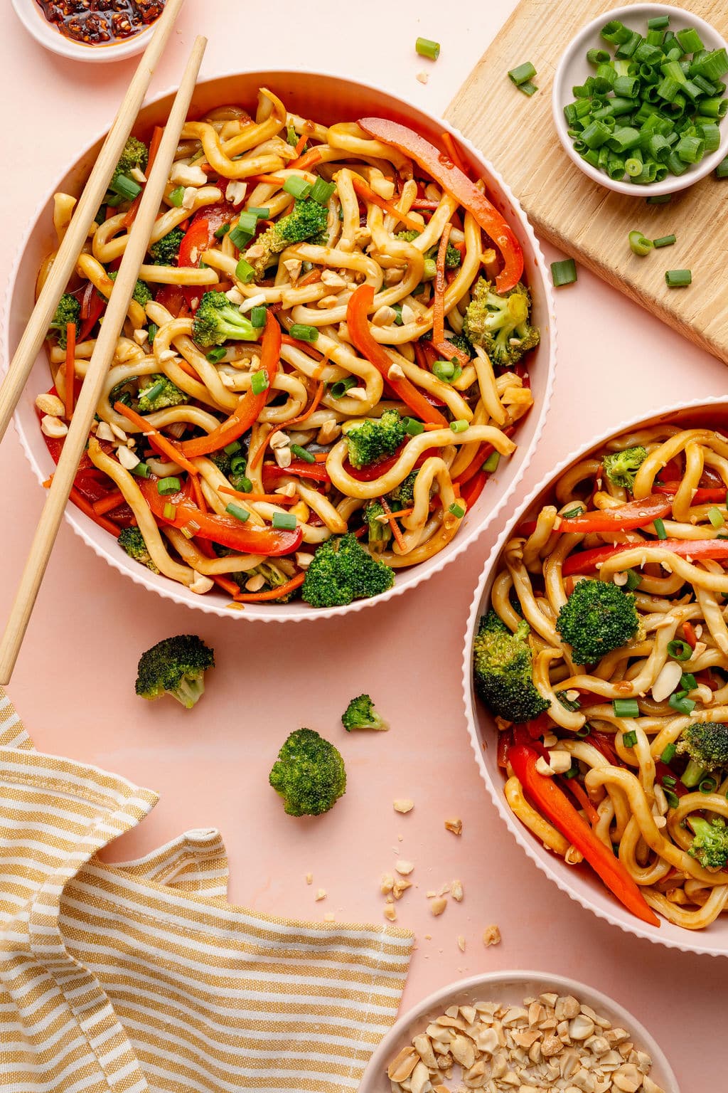 Overhead image of two bowls of sesame udon noodles tossed with broccoli, sliced red bell peppers, and carrots, garnished with green onions and chopped peanuts. Chopsticks rest in one bowl, with chili crisp, peanuts, and a striped kitchen towel on a light surface.