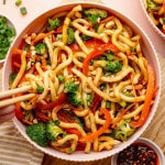 Overhead image of a bowl of sesame udon noodles with broccoli, carrots, and red bell peppers. Chopsticks lift a portion of noodles from the bowl, with chili crisp and chopped peanuts nearby.
