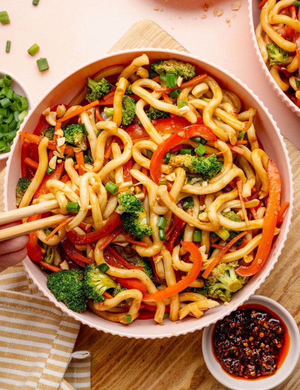 Overhead image of a bowl of sesame udon noodles with broccoli, carrots, and red bell peppers. Chopsticks lift a portion of noodles from the bowl, with chili crisp and chopped peanuts nearby.