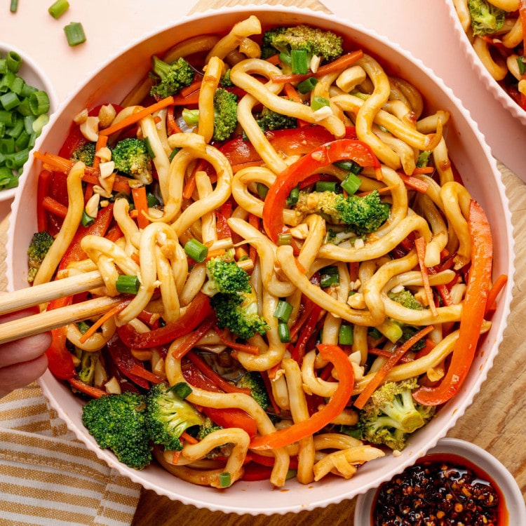 Overhead image of a bowl of sesame udon noodles with broccoli, carrots, and red bell peppers. Chopsticks lift a portion of noodles from the bowl, with chili crisp and chopped peanuts nearby.