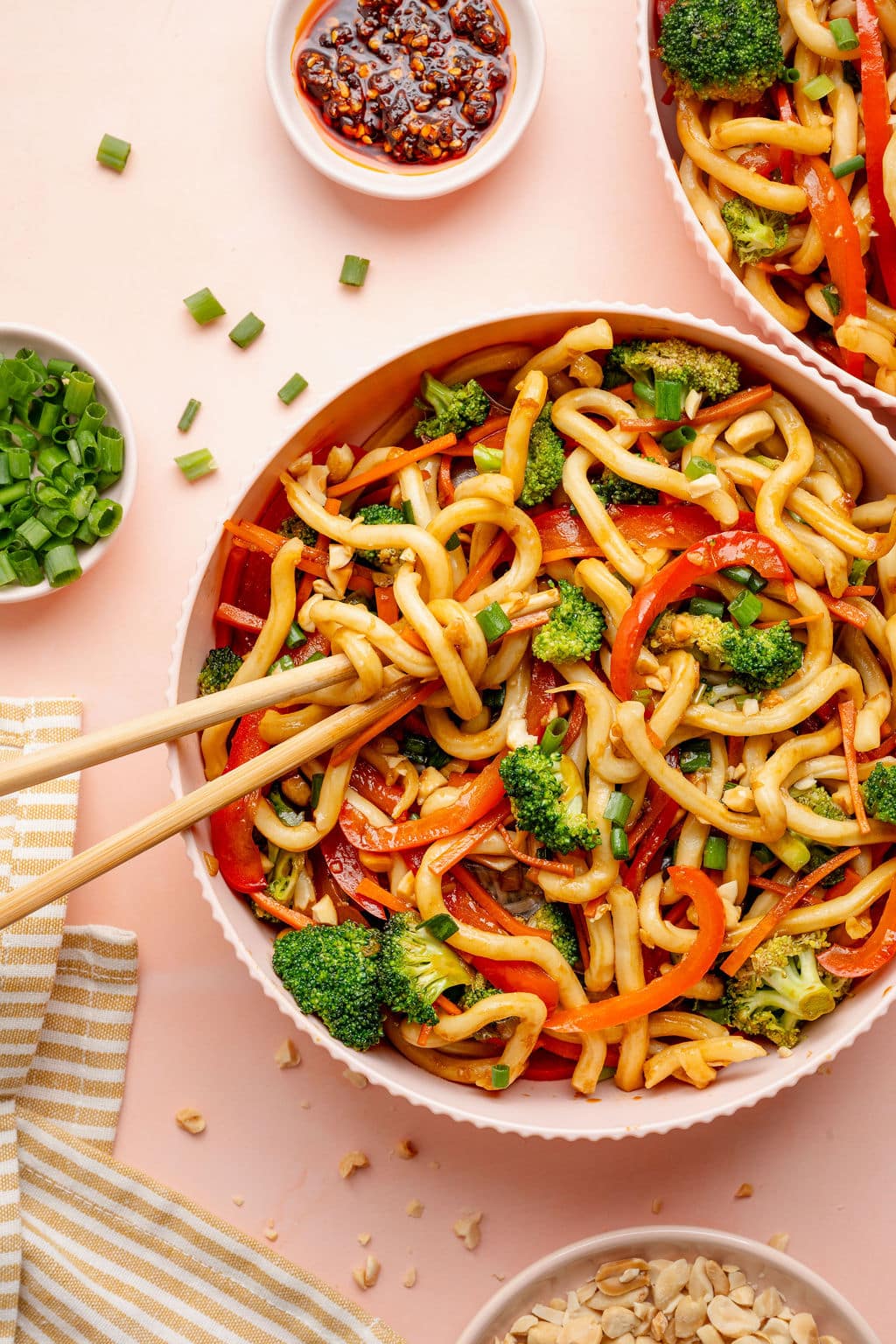 Top-down image of two bowls of sesame udon noodles with broccoli, carrots, and red bell peppers. Chopsticks lift noodles from one bowl, with small dishes of green onions, chili crisp, and chopped peanuts on the side.