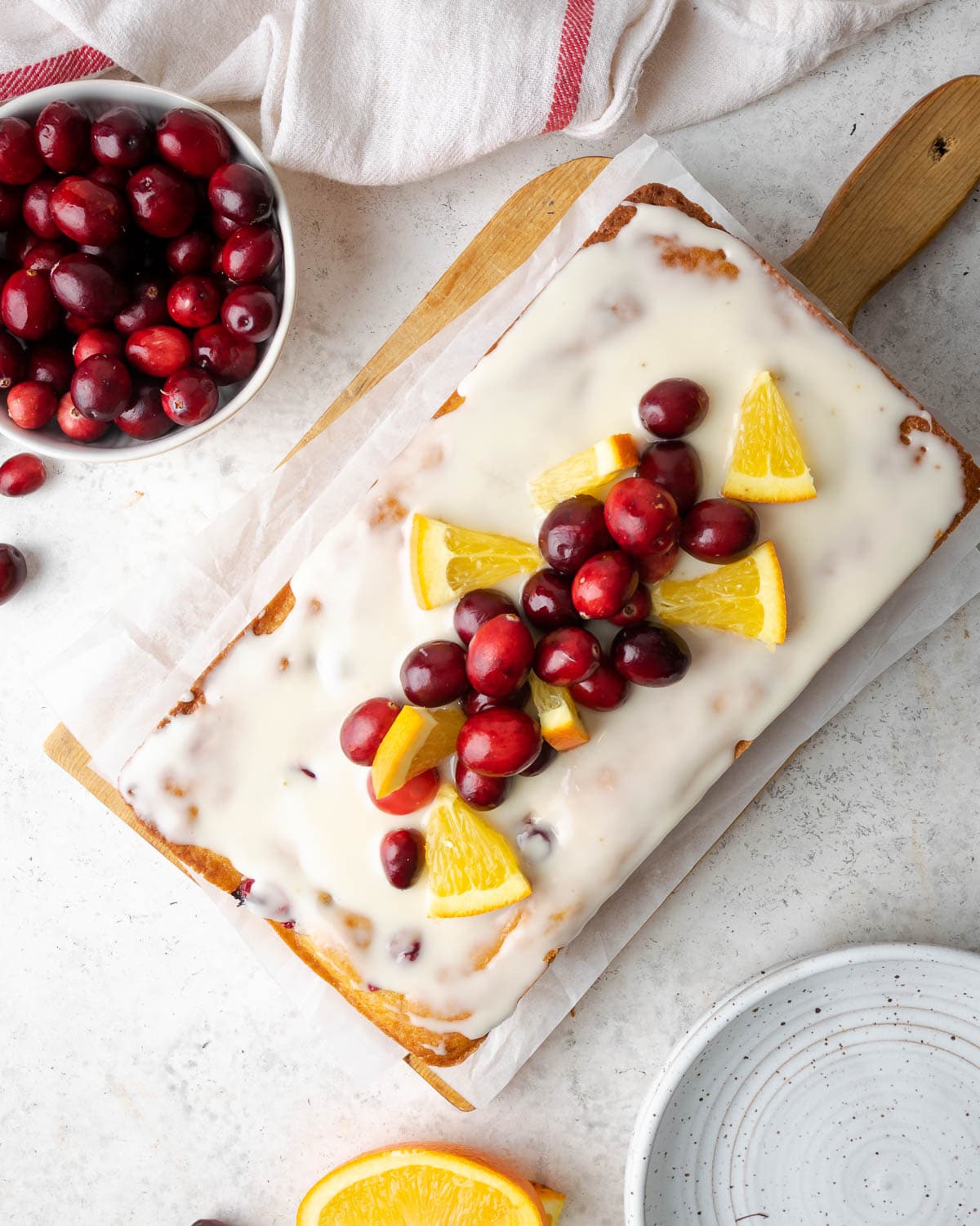 a loaf of bread on a cutting board topped with an orange glaze, fresh cranberries and oranges