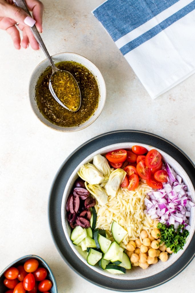 Mediterranean Orzo Salad Recipe in a large white bowl about to be tossed with Greek dressing