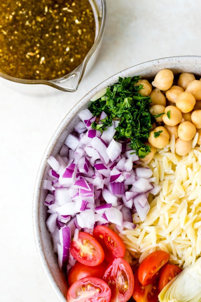 Orzo Salad Recipe in a large white bowl with red onion, cherry tomatoes, parsley and garbanzo beans