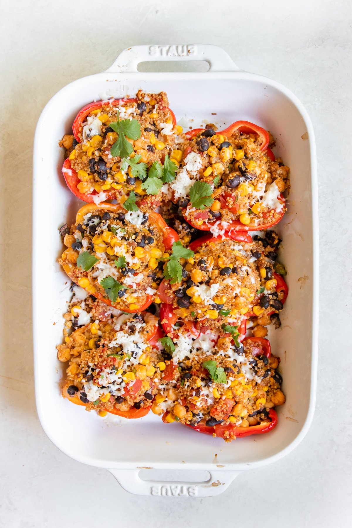 A white casserole dish with sliced bell peppers stuffed with a quiona black bean corn mixture and topped with melted cheese and cilantro