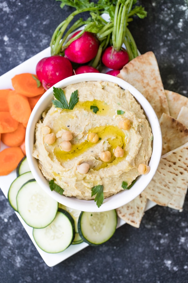 creamy homemade hummus made with tahini in a white bowl on a plate with carrots, cucumbers and pita bread 