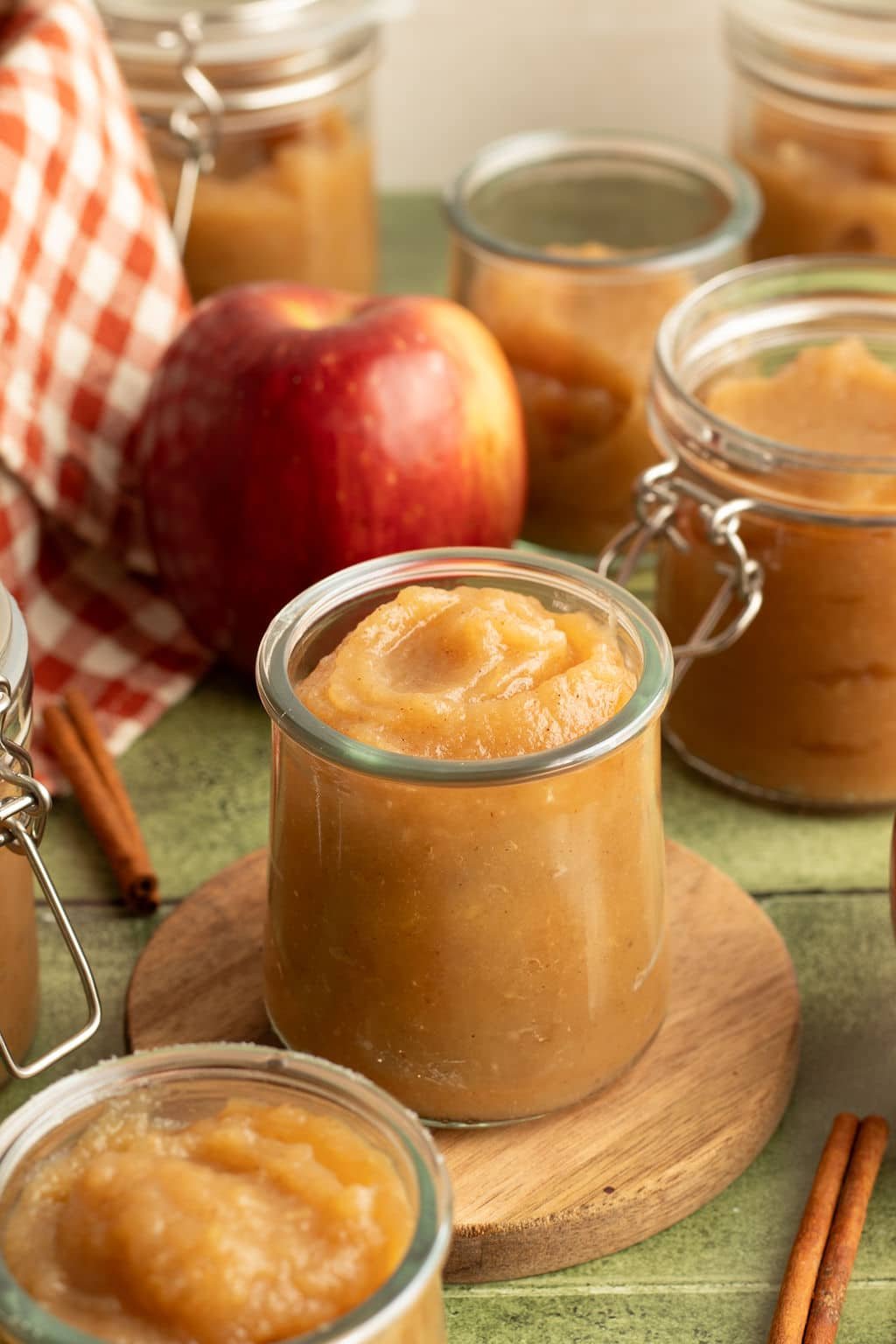Small glass jars filled with homemade applesauce on a green wooden surface, with a red apple and cinnamon sticks in the background.