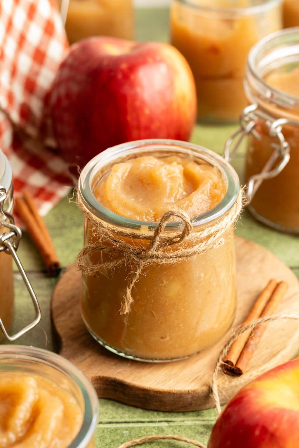 Jar of applesauce tied with rustic twine, surrounded by apples and cinnamon sticks, with more jars of applesauce in the background.