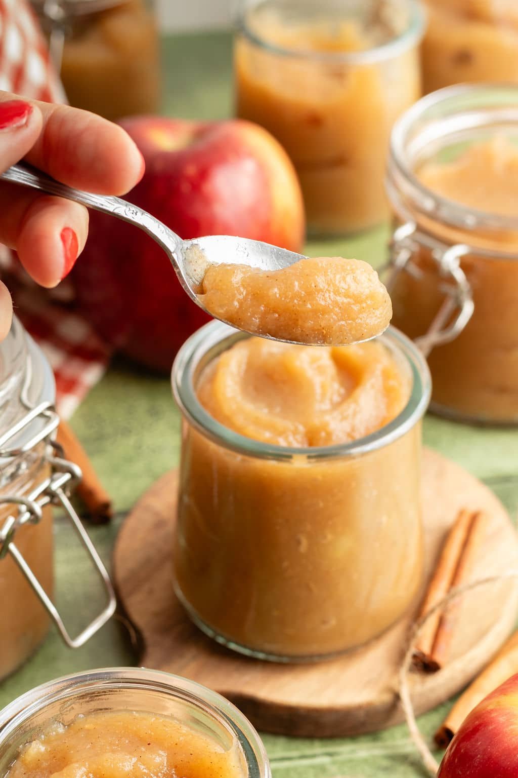 Spoonful of homemade applesauce being lifted from a glass jar, with apples and jars of applesauce in the background.