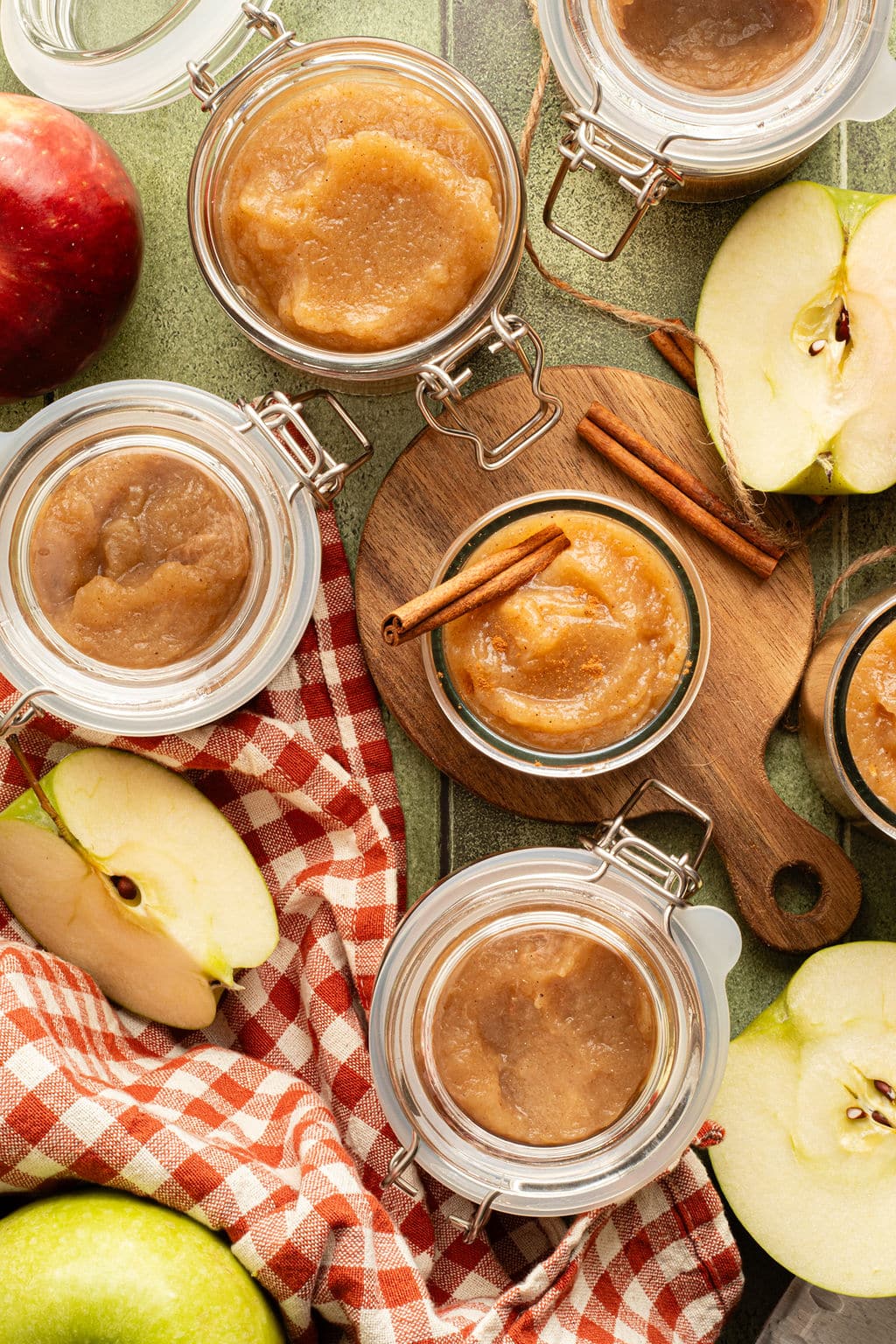 Overhead view of multiple jars of homemade applesauce on a green table with apples, a red checkered cloth, and cinnamon sticks for garnish.