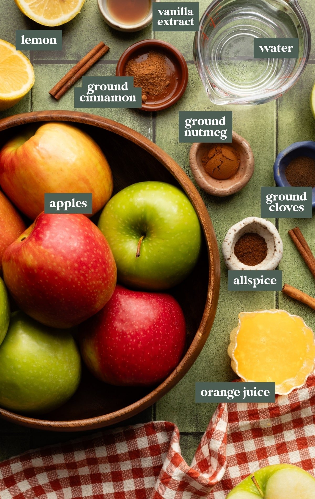 Overhead view of homemade applesauce ingredients on a green tiled surface with a red gingham napkin, including a wooden bowl filled with fresh red and green apples, halved lemon, small bowls of ground cinnamon, ground nutmeg, ground cloves, and allspice, a measuring cup of water, vanilla extract, orange juice, and cinnamon sticks.