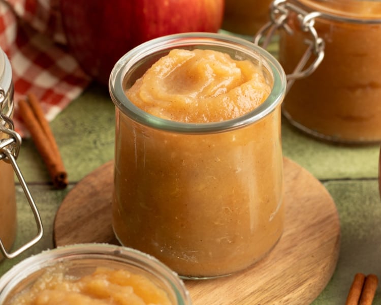 Jar of homemade applesauce placed on a wooden coaster, surrounded by other jars, cinnamon sticks, and a fresh apple in the background.