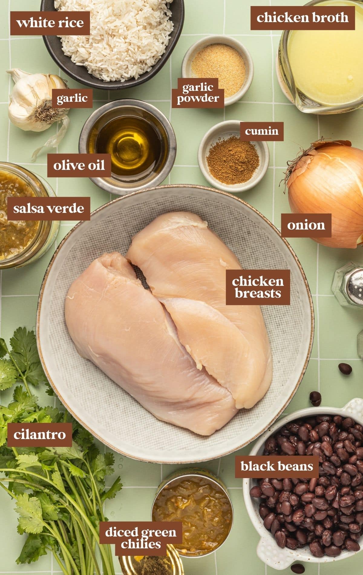 Ingredients in bowls with text labels on them. Includes white rice, chicken brother, garlic powder, garlic, cumin, olive oil, onion, salsa verde, raw chicken, cilantro, green chilies, and black beans.