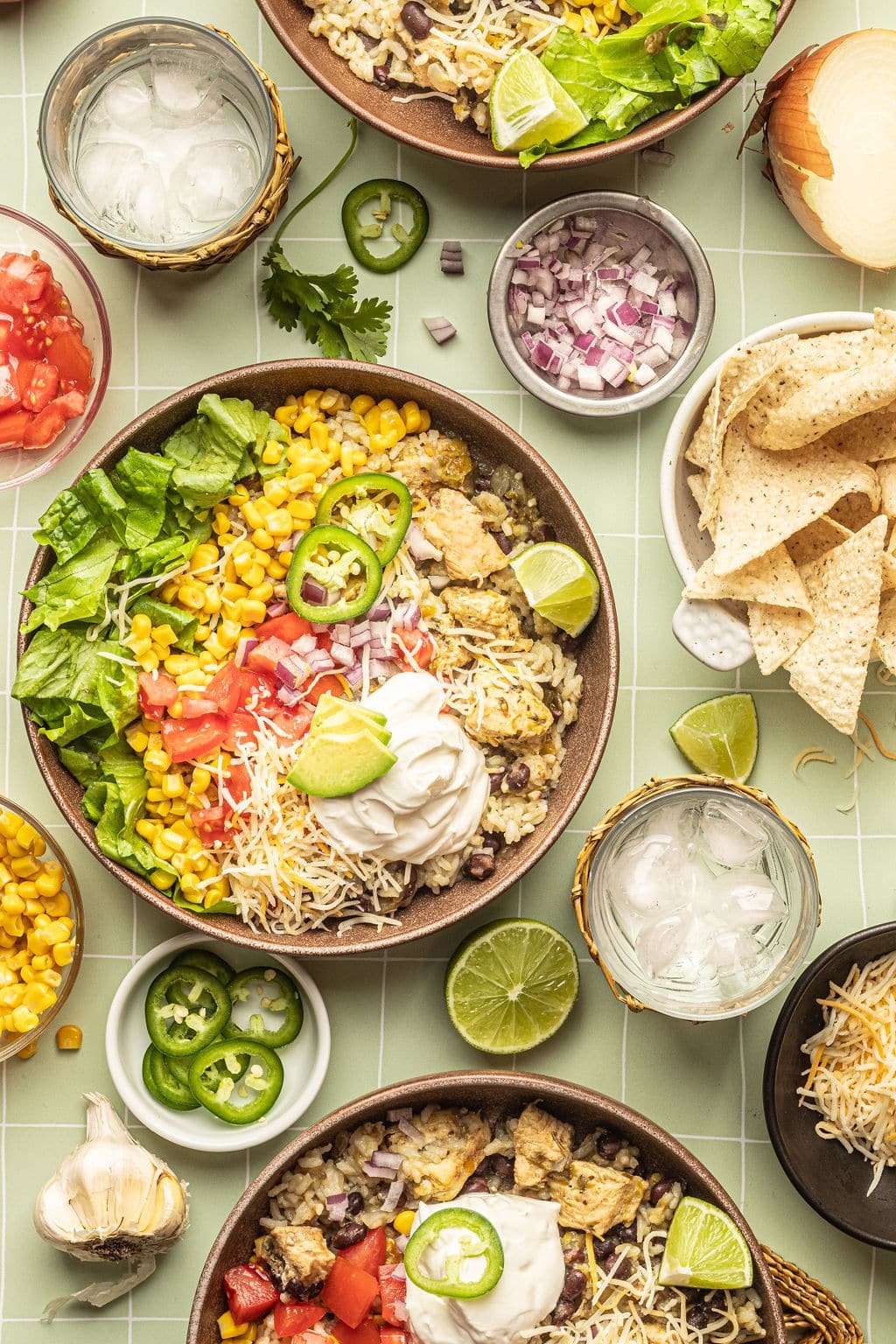 A bowl with white rice, chicken, black beans, cheese, corn, lettuce, tomatoes, diced onions, sour cream, lime wedges, and sliced jalapeno. Surrounded by tortilla chips, bowl of diced red onion, bowl with sliced jalapeno, lemon wedges, and a glass of ice water.