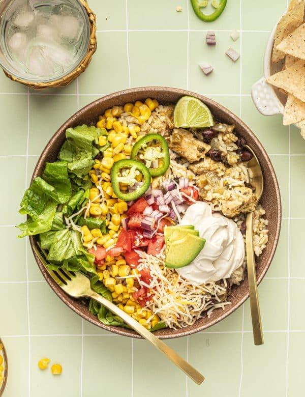 A bowl with a gold fork and spoon and white rice, chicken, black beans, cheese, corn, lettuce, tomatoes, diced onions, sour cream, lime wedges, and sliced jalapeno.